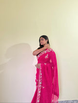 Woman wearing a pink saree with white patterns against a plain wall.